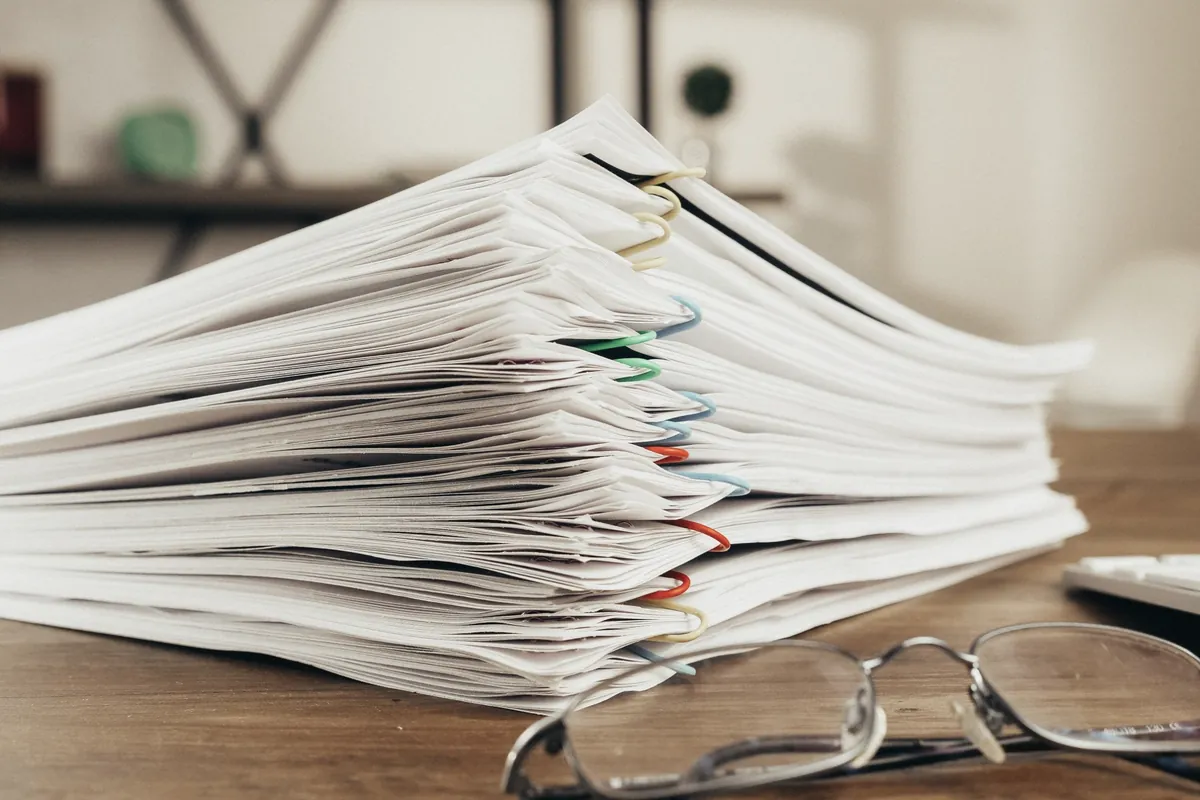 Pile de documents attachés par des trombones sur un bureau en bois, à côté d'une paire de lunettes et d'un clavier